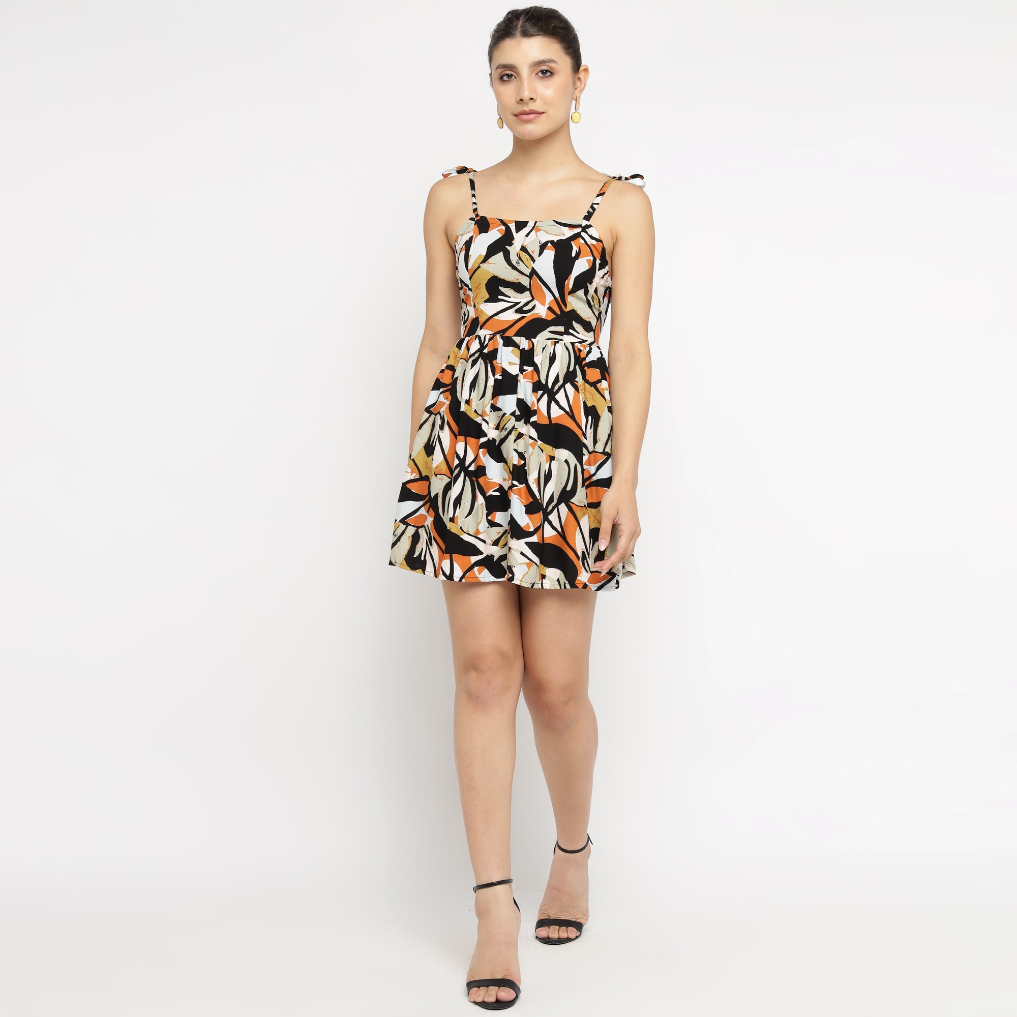 Woman wearing a tropical leaf print mini dress with tie-up shoulder straps, front view showing flared silhouette and above-knee hem, paired with black heels.