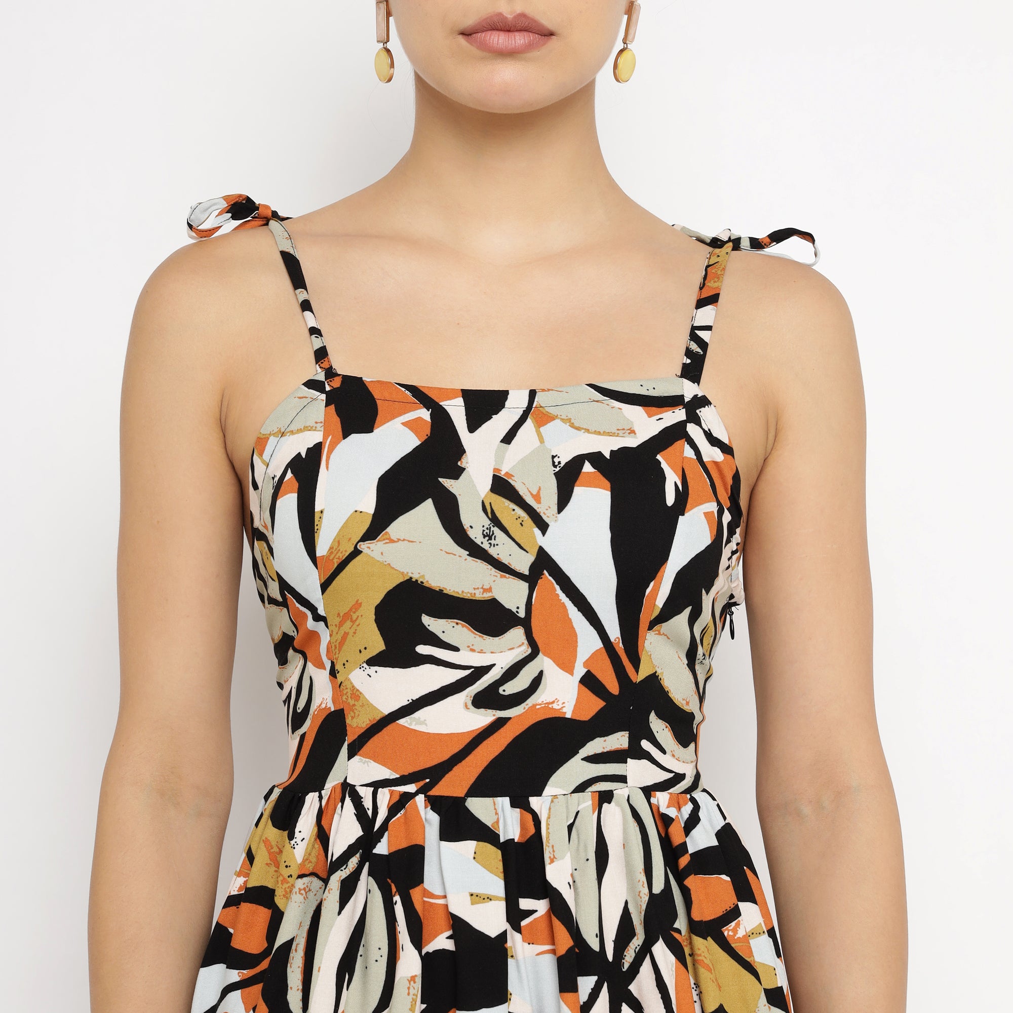 Front view of a woman wearing a tropical leaf print strappy mini dress with tied shoulder straps, featuring a fitted bodice and flared waist in black, orange, and beige tones.