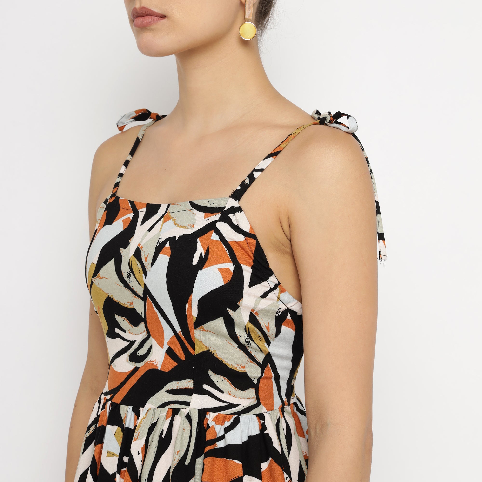Close-up side view of a woman wearing a tropical leaf print mini dress with tie-up spaghetti straps in black, orange, and beige tones.