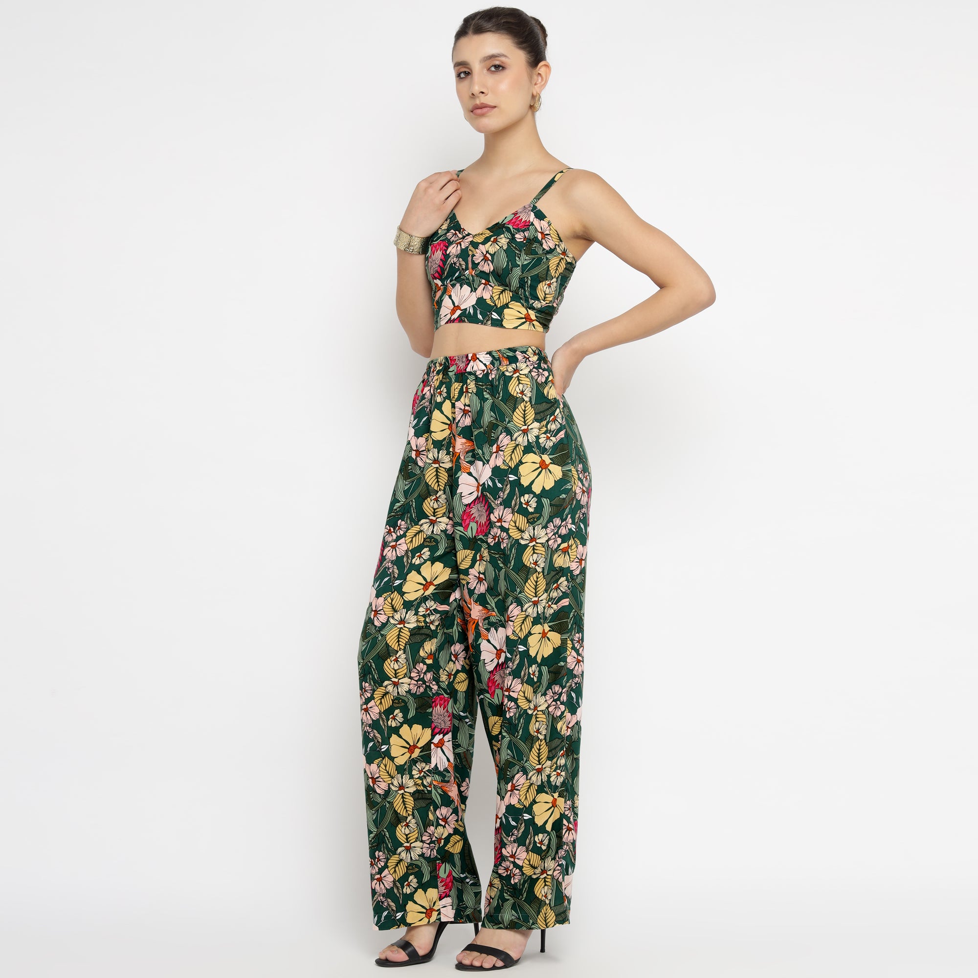 Full-length view of a model wearing the Tropical Bloom Co-ord Set, which features a sleeveless crop top with thin straps and high-waisted wide-leg pants. The outfit displays a vibrant tropical floral print in green, yellow, pink, and red tones. The model poses confidently with one hand on her hip and the other near her shoulder, accessorized with earrings, a bracelet, and black heels.