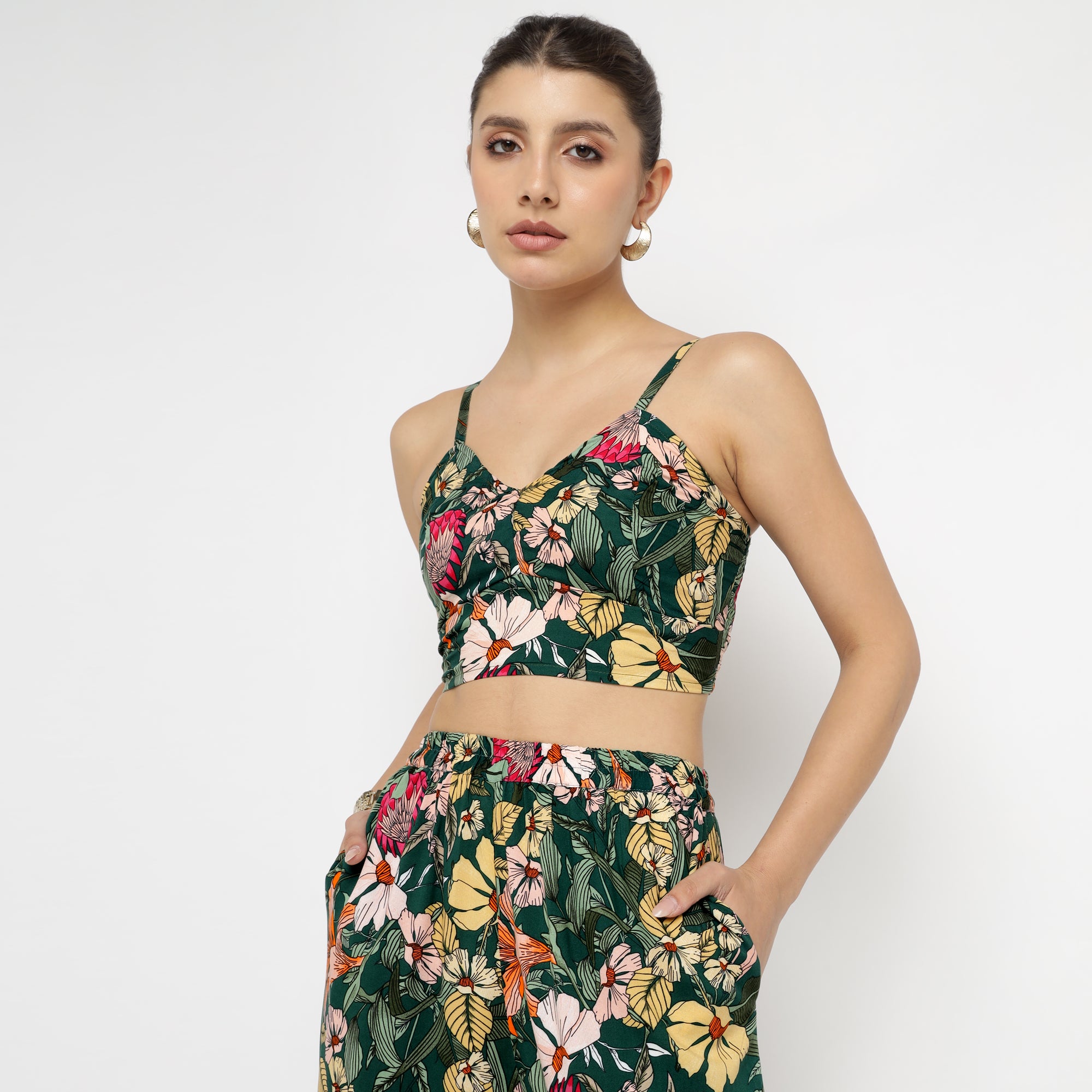 Model wearing the Tropical Bloom Co-ord Set, featuring a coordinated sleeveless crop top and high-waisted pants. The outfit has a vibrant tropical floral print with colors like yellow, red, pink, and white on a deep green background. The top has thin straps and a V-neckline, while the pants have side pockets and a comfortable elastic waistband. The model poses confidently with one hand on her hip.